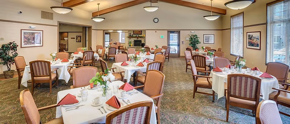 Spacious dining room with multiple round tables set with white linens, folded napkins, floral centerpieces and chairs.