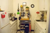 Small galley kitchen with white cabinets, a sink and refrigerator, and a central metal shelving rack holding utensils and dishes.