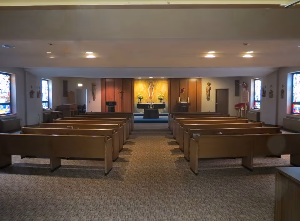 Interior view of a chapel with wooden pews arranged in rows facing an altar adorned with religious statues and flowers. Stained glass windows line the side walls, and the room is softly lit by ceiling lights.