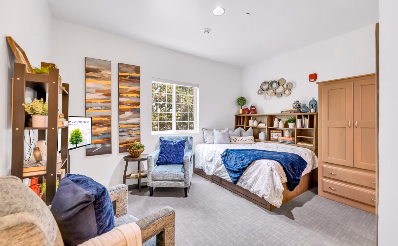 A bright and cozy bedroom in a senior living community featuring a bed with white and blue bedding, a wooden headboard with shelves holding decorative items and books, two comfortable armchairs with blue pillows, a small round side table with a plant, a wooden cabinet, and two abstract paintings on the wall next to a window letting in natural light.