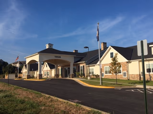 Exterior view of Lansdowne Heights facility showing a single-story building with a covered entrance, a driveway, and two flagpoles with flags. The building has beige siding with stone accents and multiple windows under a dark roof, set against a blue sky.