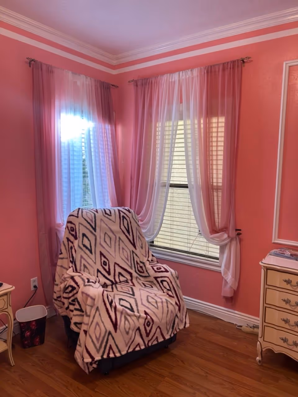 A cozy corner of a room with pink walls and two windows covered with sheer white and pink curtains. There is a comfortable armchair draped with a patterned blanket in front of the windows. To the right of the chair is a small cream-colored dresser with decorative handles, and to the left is a small side table with a trash bin underneath. The floor is wooden.