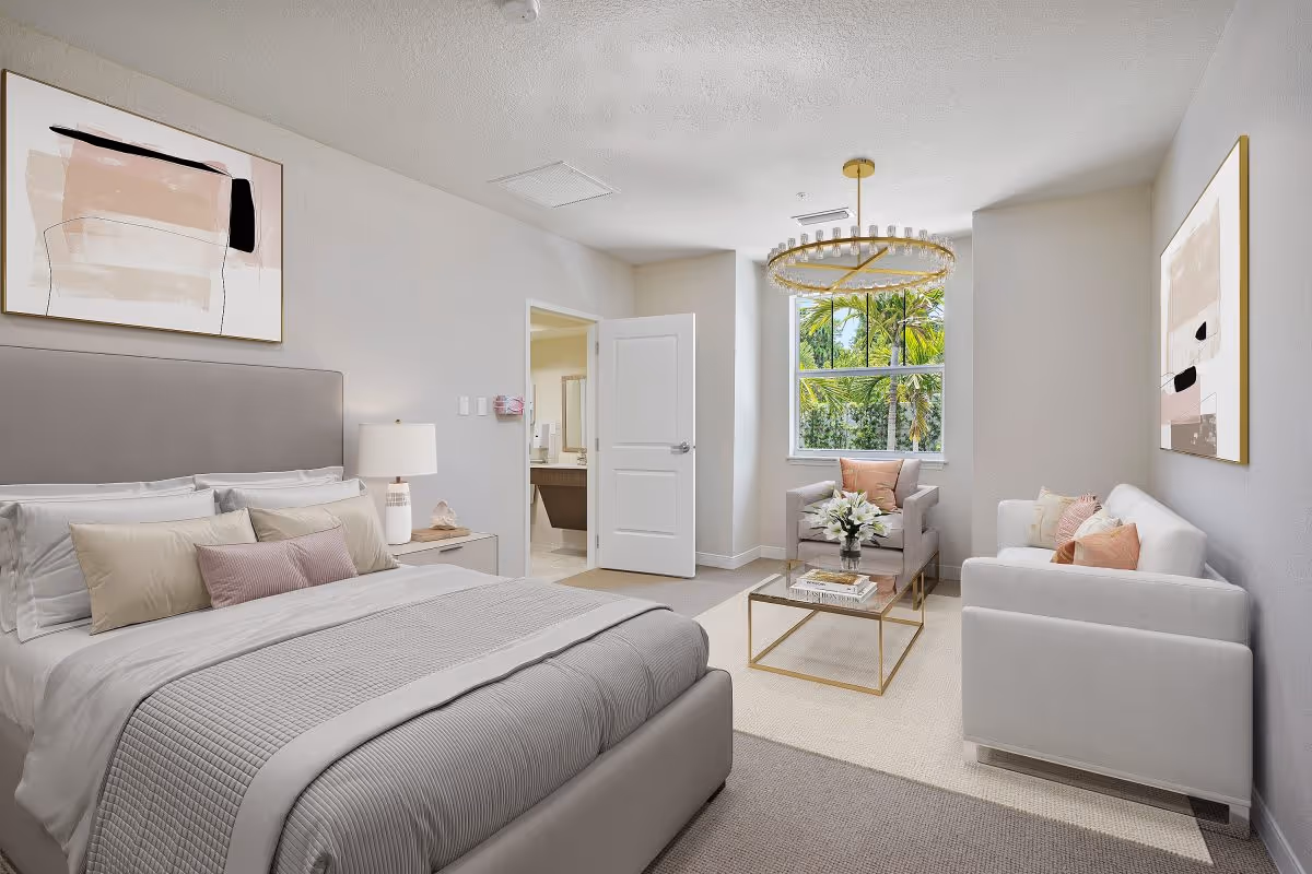 Bright furnished bedroom with a bed, a seating area including a sofa and armchair, a coffee table, and a window showing greenery.