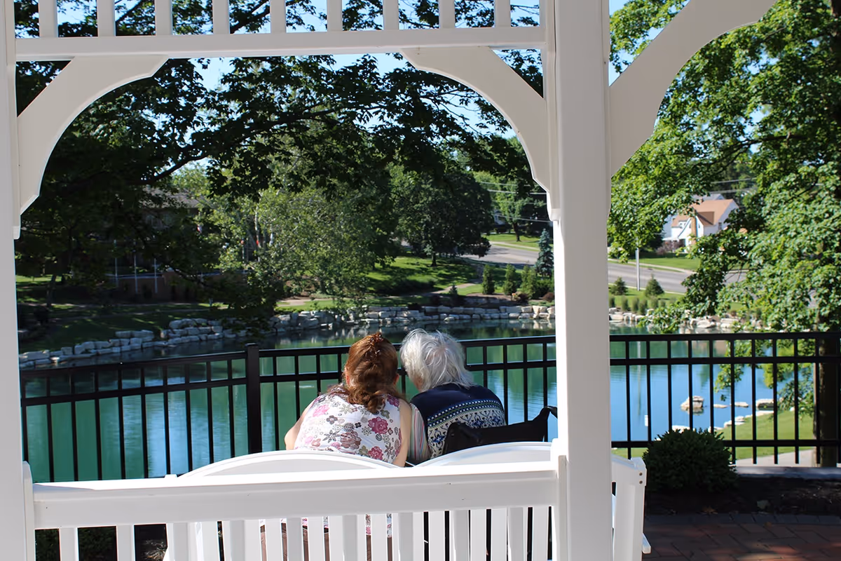Two people sitting on a white bench under a white pergola, overlooking a serene pond with trees and houses in the background on a sunny day.