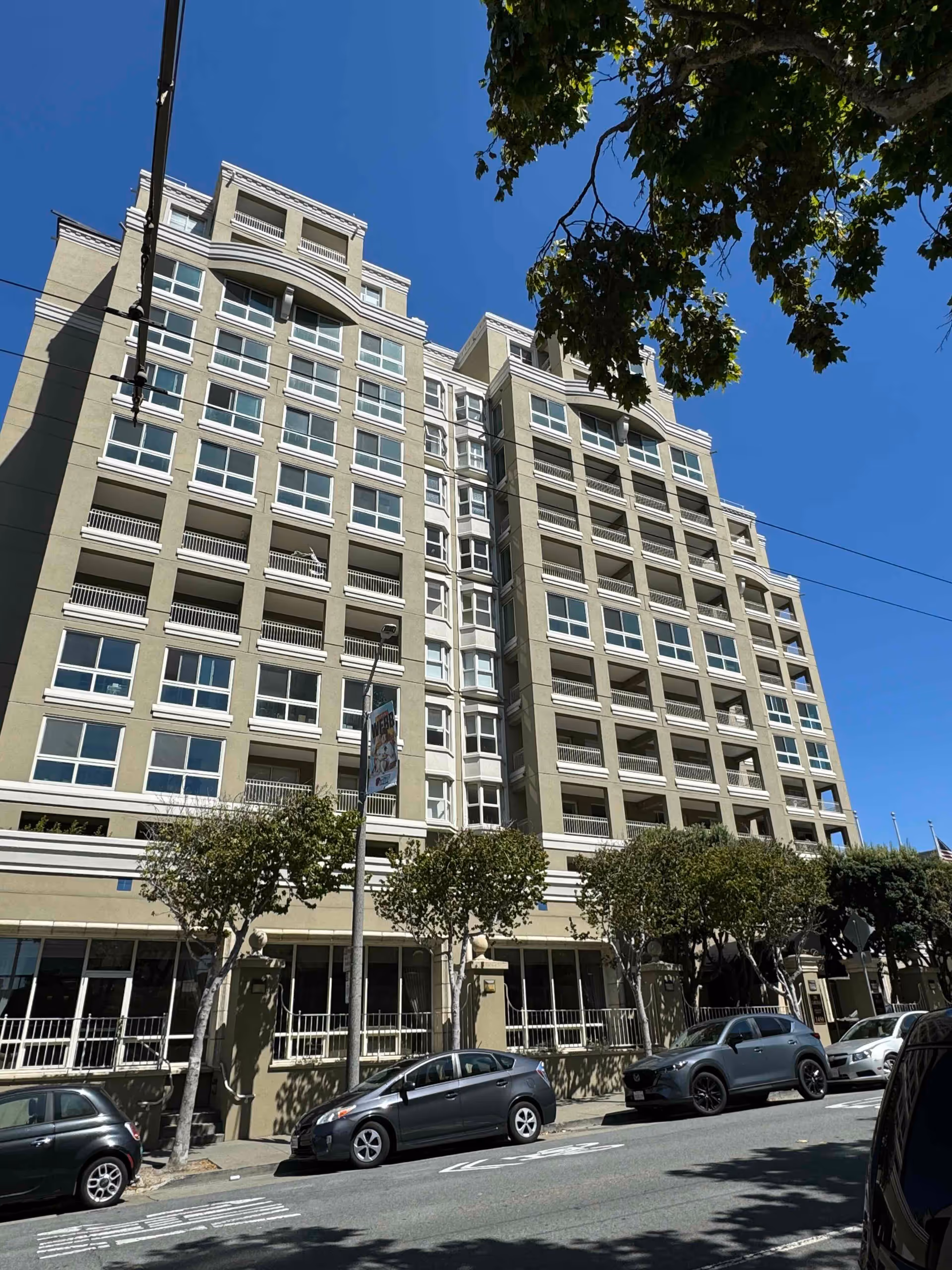 Exterior view of a multi-story residential building with numerous windows and balconies under a clear blue sky. Several cars are parked along the street in front of the building, and trees line the sidewalk.