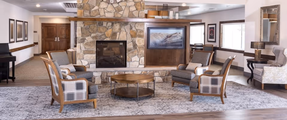 A cozy living room area featuring a stone fireplace in the center with a wooden mantel. Four cushioned armchairs with wooden frames and plaid upholstery are arranged around a round wooden coffee table on a large patterned rug. The room has wooden flooring, a piano on the left side, framed artwork on the walls, and a window with blinds on the right side. A side table with a lamp and a decorative chair are also visible.