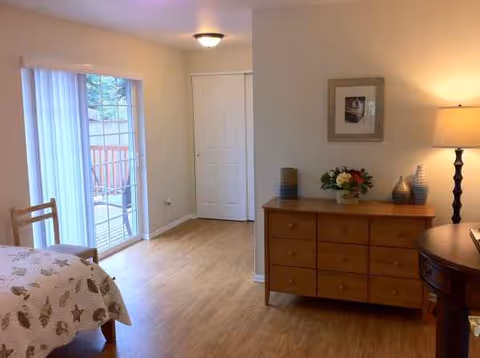 Bright bedroom with bed on left, sliding doors to balcony, and wooden dresser against wall.