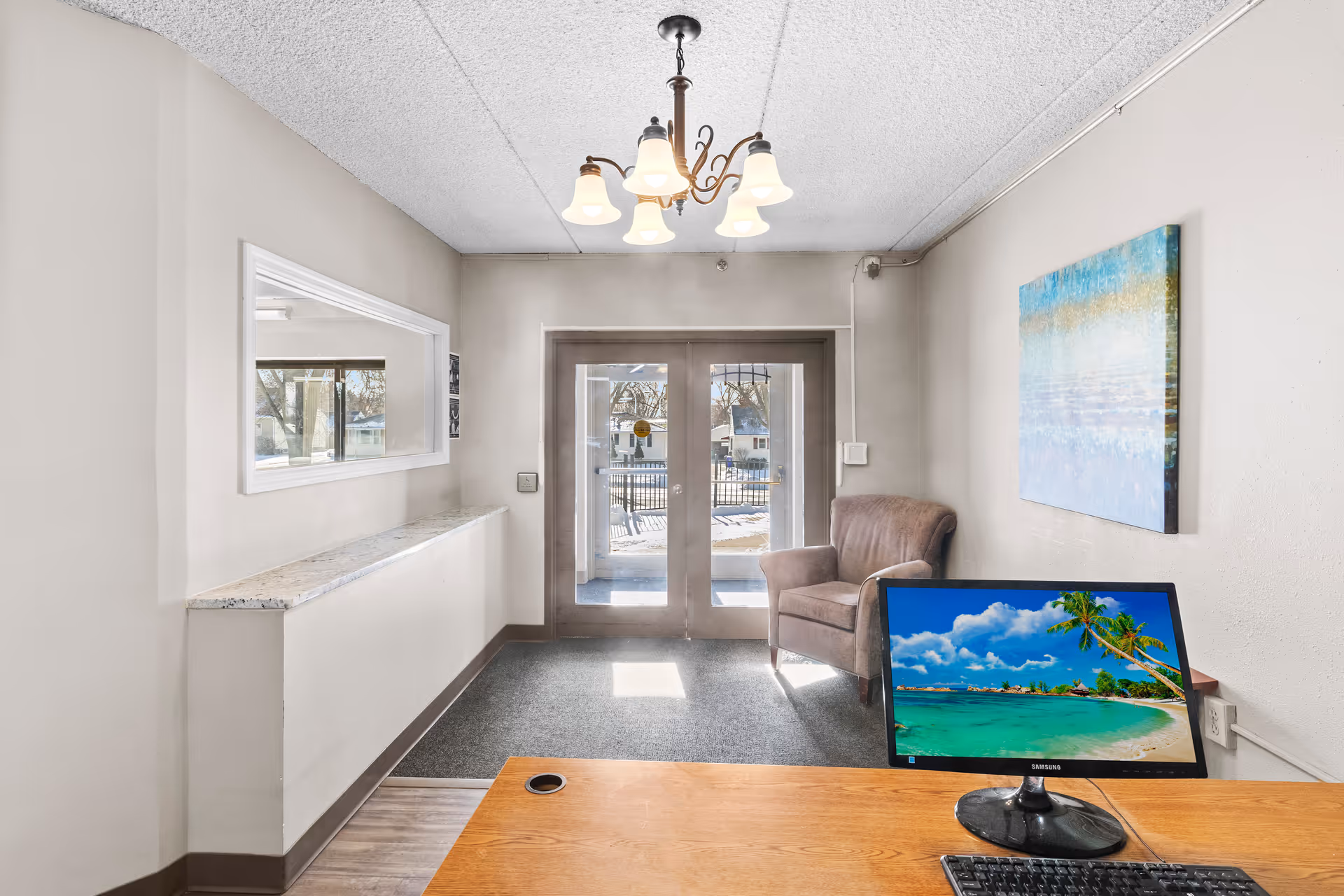 Reception area with a wooden desk featuring a computer monitor displaying a tropical beach scene. Behind the desk is a brown armchair next to double glass doors leading outside. A chandelier with six lights hangs from the ceiling, and a colorful abstract painting is on the wall to the right. A large window opening is on the left wall.