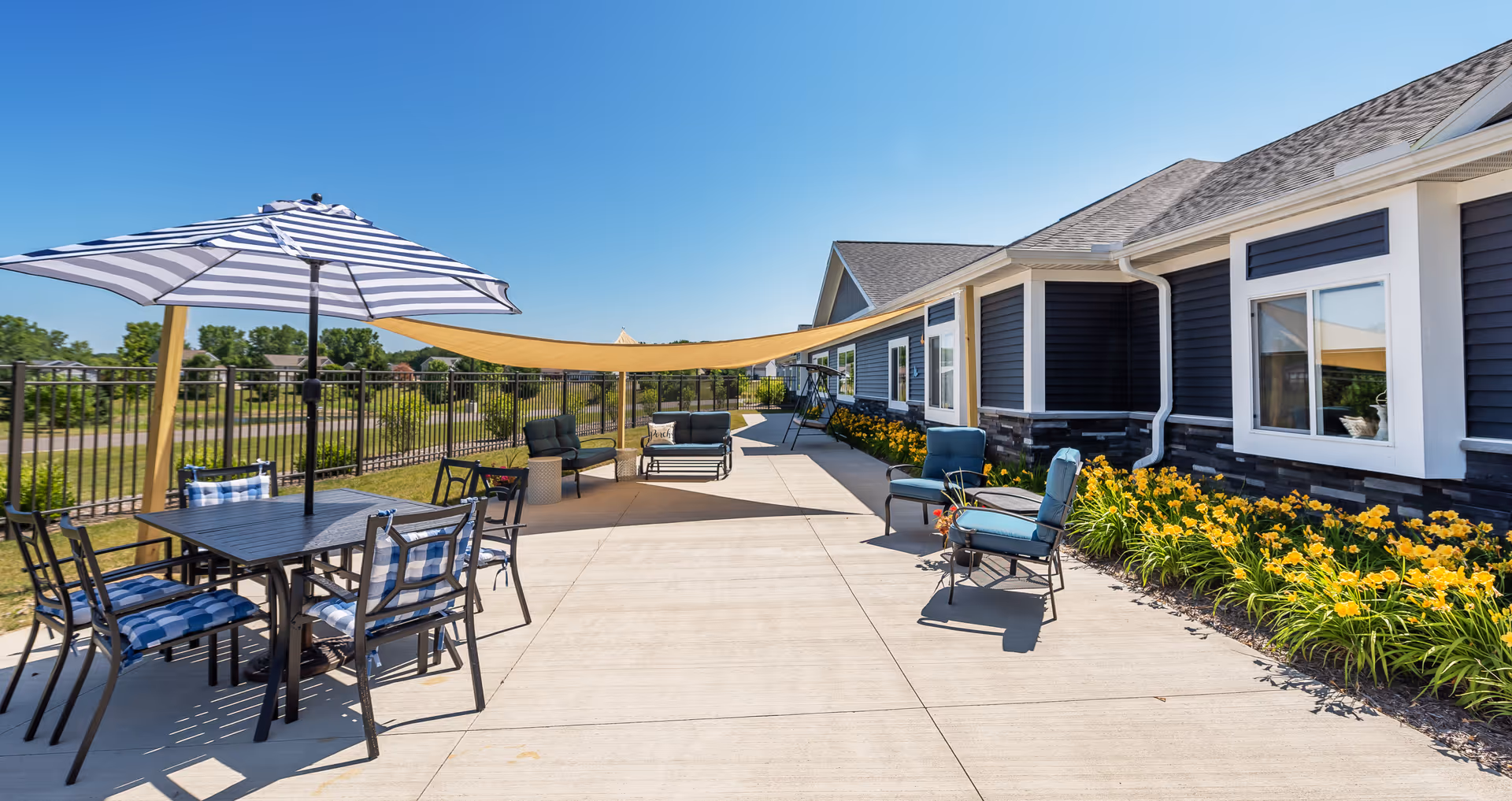 Outdoor patio with tables, chairs, umbrellas, shade sails and flowerbeds along the side of a senior living building.