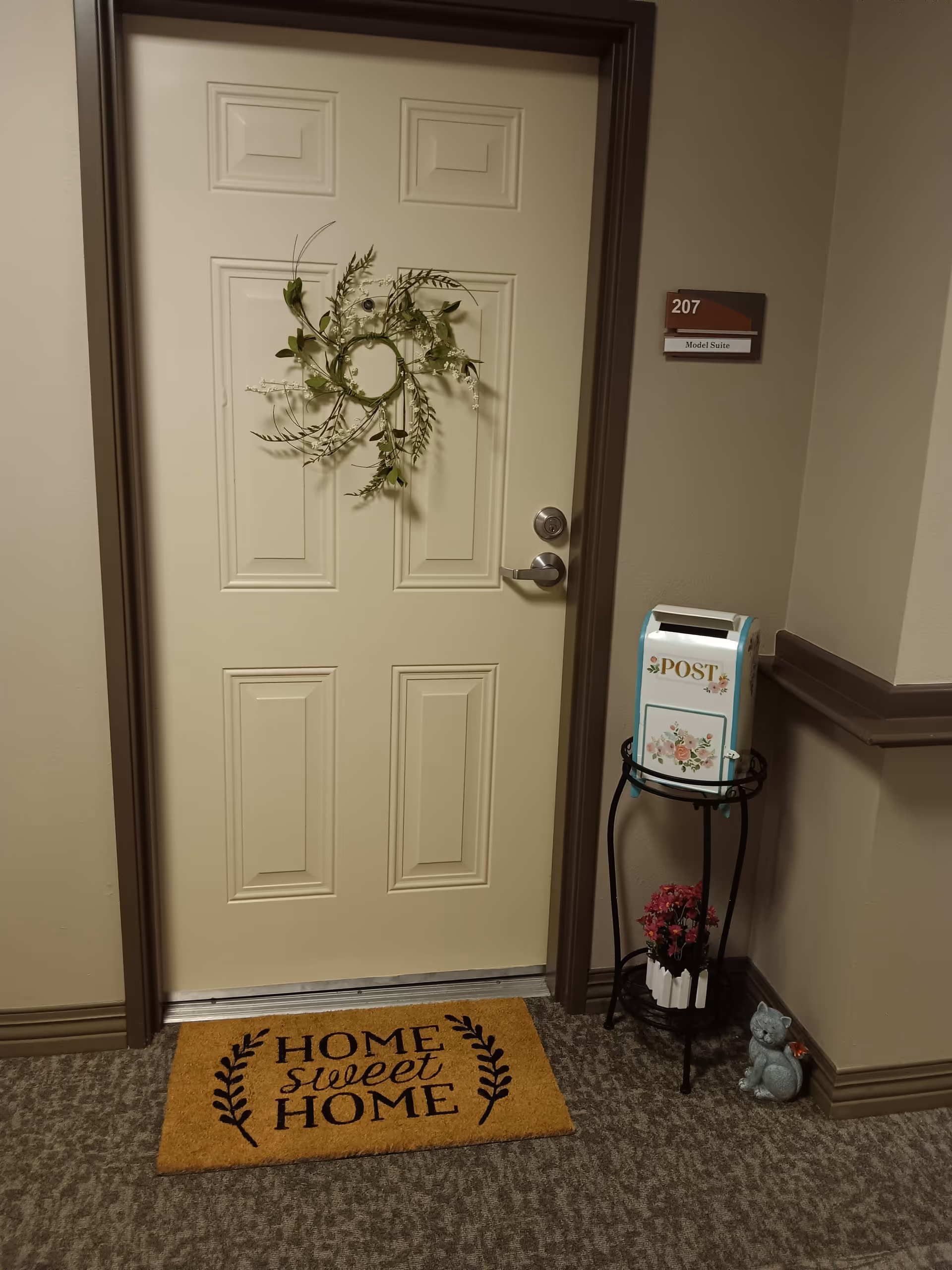 A closed beige door with a decorative green wreath hanging on it. The door has a silver handle and a peephole. To the right of the door is a small stand holding a decorative mailbox labeled 'POST' with floral designs and a small flower pot with pink flowers underneath. A small cat figurine is placed on the floor next to the stand. A brown doormat in front of the door reads 'HOME sweet HOME'. A sign on the wall next to the door reads '207 Model Suite'.