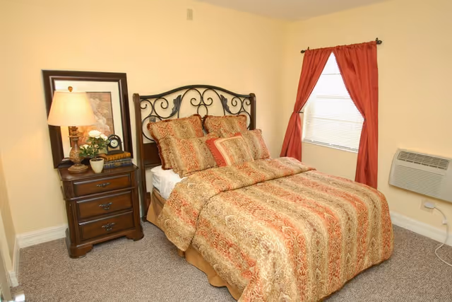 A cozy bedroom with a double bed featuring an ornate metal headboard and a patterned bedspread in shades of gold, red, and beige. Next to the bed is a wooden nightstand with three drawers, a table lamp, a framed picture, a small clock, and a potted plant. The room has beige walls, a window with red curtains, and a wall-mounted air conditioning unit below the window.