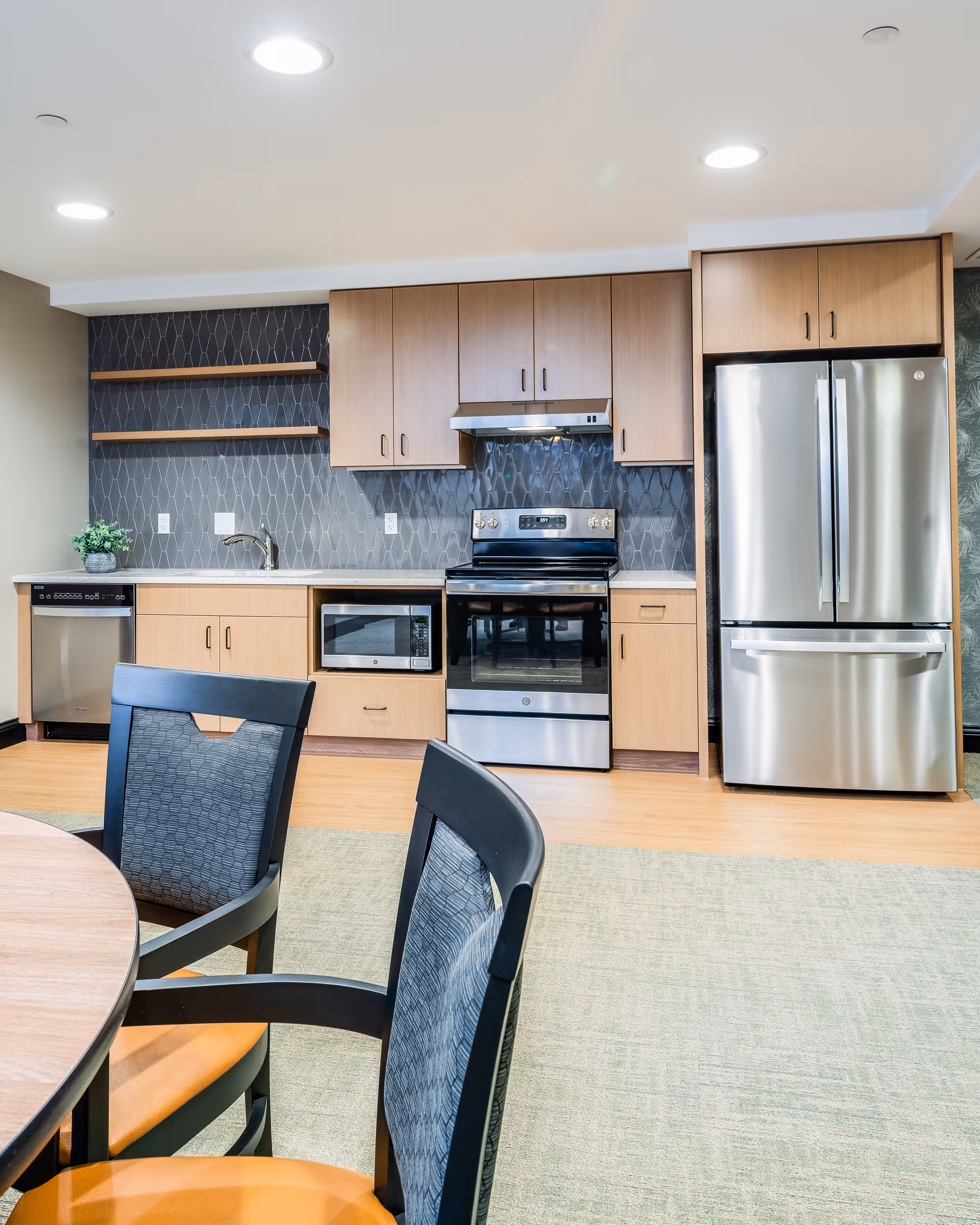 Modern communal kitchen with stainless steel appliances, light wood cabinets, and a round dining table with chairs in the foreground.