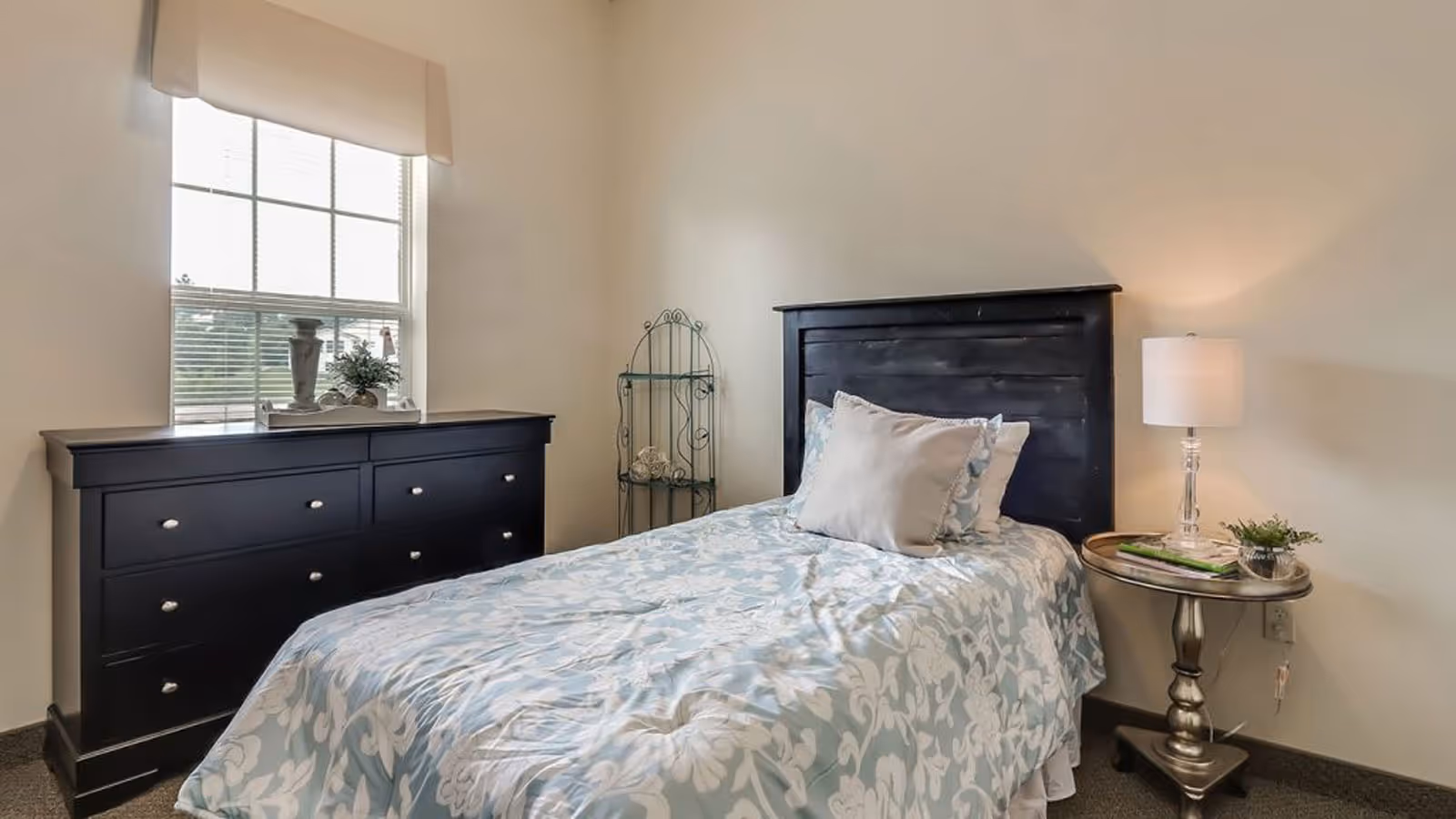 A small bedroom with a single bed featuring a black wooden headboard and floral patterned bedding in light blue and white. There is a black dresser under a window with blinds and a beige valance. Next to the bed is a round metallic side table with a lamp, a small plant, and some magazines. A decorative metal shelf stands in the corner.