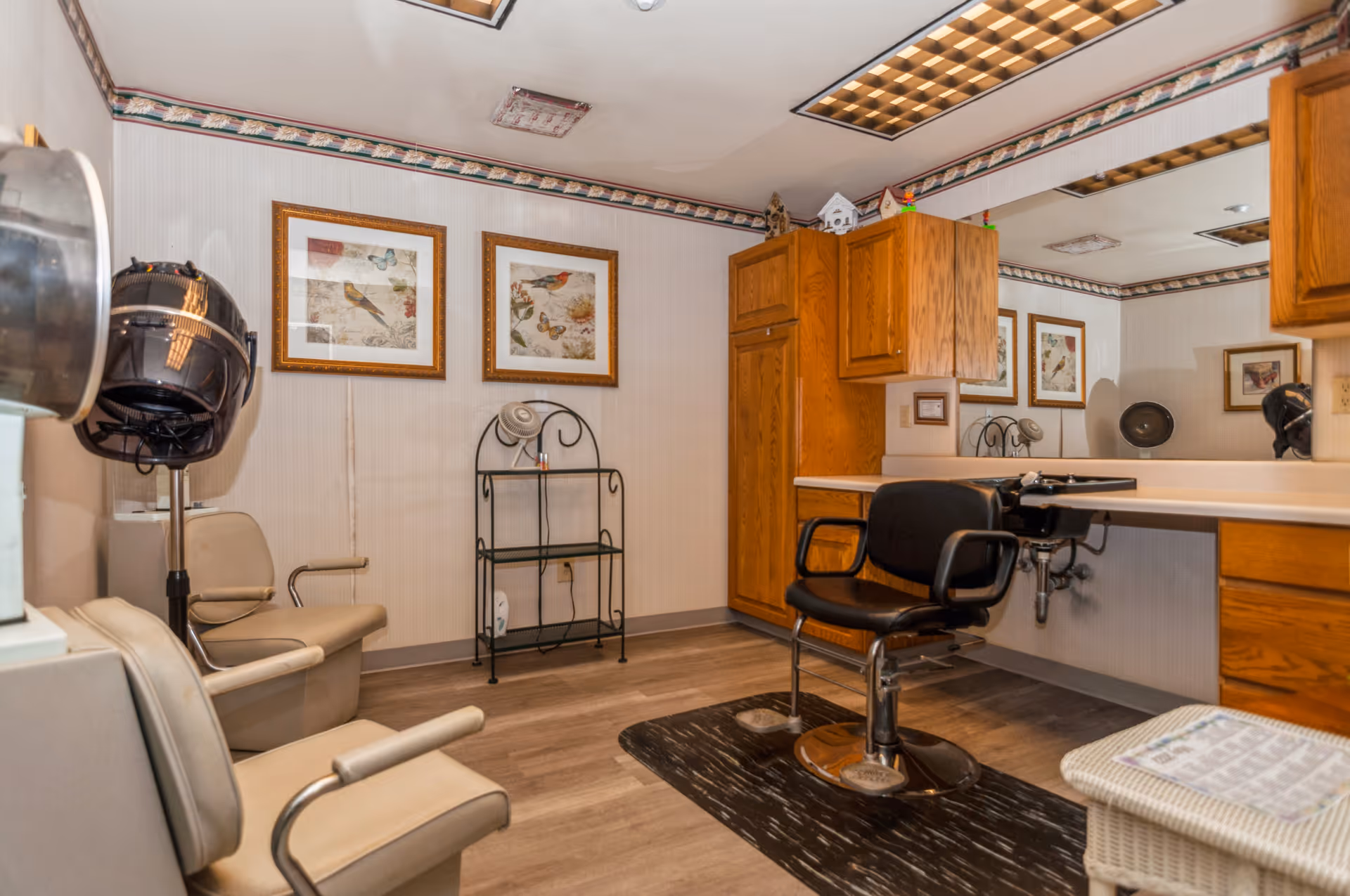 A senior living salon room with a styling chair, hooded hair dryer, mirror, and wooden cabinets.