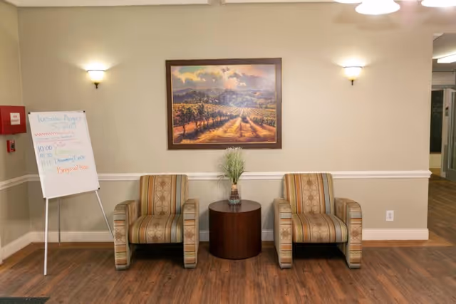 Two upholstered chairs flank a round wooden table topped with a vase beneath a framed landscape painting in a reception/lounge area with a whiteboard on an easel to the left.