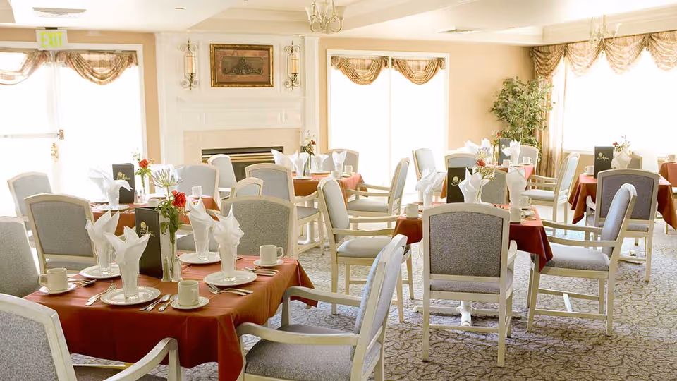 Bright dining room with multiple tables set with white napkins, plates and cups, upholstered chairs and a fireplace.