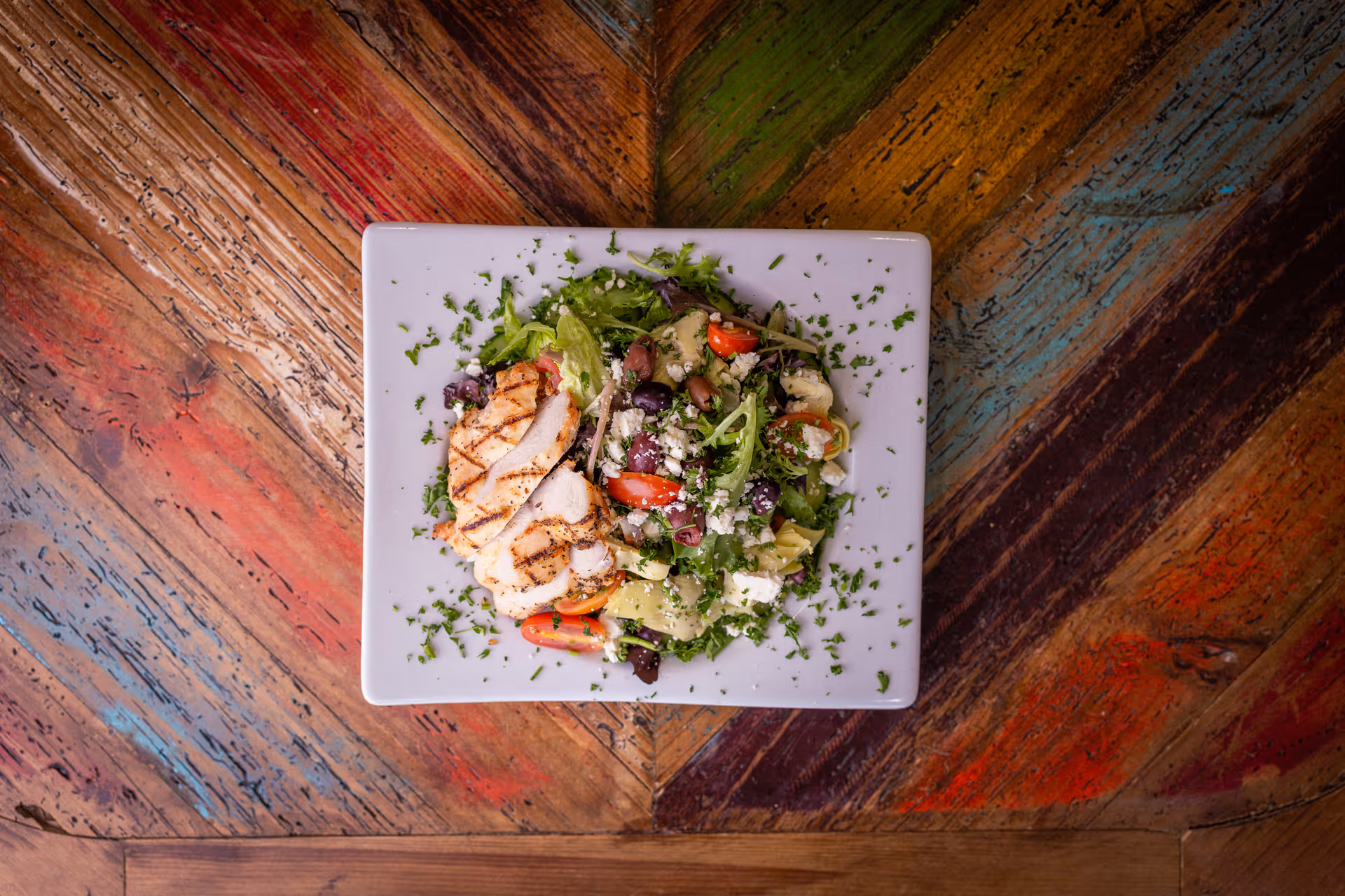 A white square plate with a grilled chicken breast and a mixed salad containing lettuce, cherry tomatoes, olives, artichoke hearts, and crumbled cheese, garnished with chopped herbs, placed on a colorful wooden table with a chevron pattern.