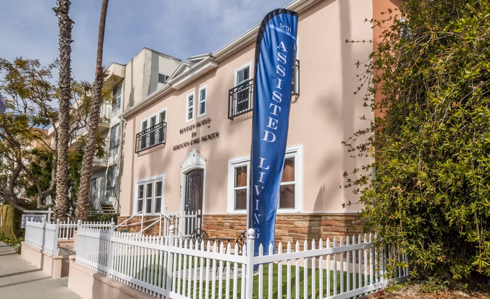 Exterior view of a two-story assisted living facility named Bentley Suites by SCH, featuring a light pink facade with white trim, small balconies, a white picket fence, and a tall blue vertical banner that reads 'ASSISTED LIVING'. There are palm trees and other greenery around the building under a partly cloudy sky.
