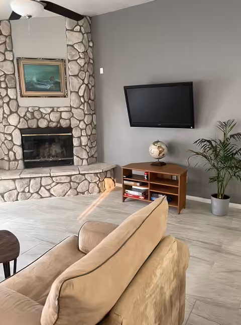 A living room with a stone fireplace on the left wall, a mounted flat-screen TV on the right wall above a wooden TV stand with a globe and some books. There is a beige couch in the foreground and a potted plant in the corner near the TV stand. The floor is light-colored tile and the walls are painted gray.
