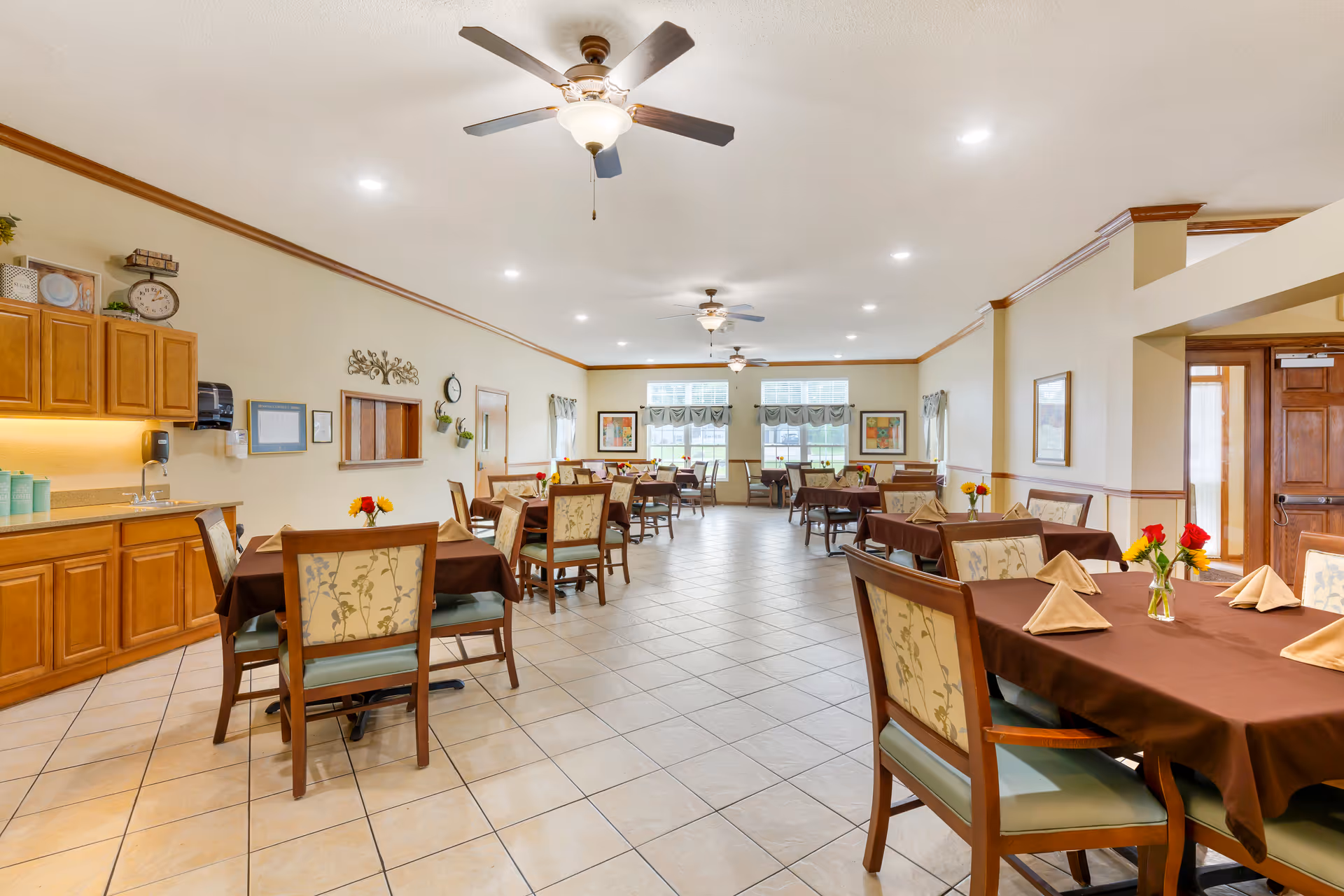 A spacious dining room with multiple tables covered in brown tablecloths, each set with beige folded napkins and small flower vases. The room has tiled floors, beige walls with wooden trim, ceiling fans with lights, and large windows letting in natural light. There is a counter with cabinets and a sink on the left side of the room.