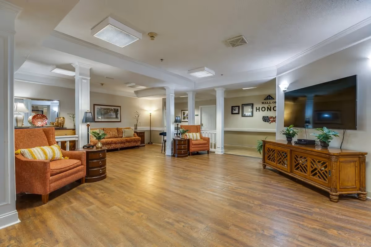 Spacious senior living common lounge with armchairs, sofas, columns, wood flooring and a large wall-mounted TV.