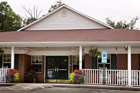 Front exterior view of a single-story brick building with a red roof and white trim. The entrance features double glass doors with green frames, flanked by windows with green shutters. There are hanging plants and potted flowers near the entrance, a white railing along the porch, and a handicapped parking sign in front.