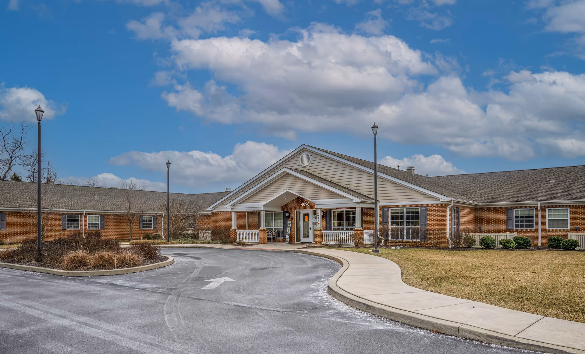 Exterior view of a single-story brick building with a covered entrance and a curved driveway. The building has multiple windows and is surrounded by a lawn and some landscaping. The sky is partly cloudy with blue patches visible.