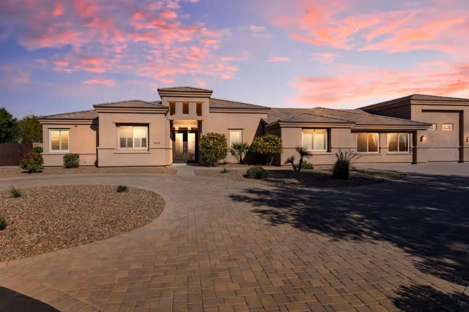 Exterior view of a single-story residential building with a paved driveway and desert landscaping under a colorful sunset sky.
