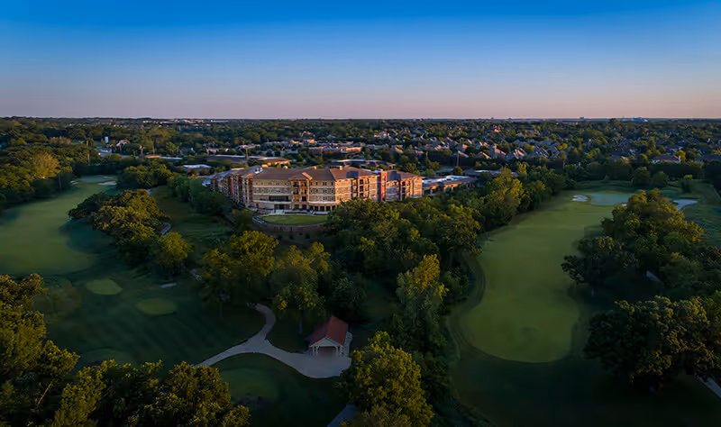 Aerial view of the Fairways of Ironhorse Independent Living facility surrounded by lush green trees and adjacent to a golf course with well-maintained fairways and putting greens, under a clear sky during sunset.