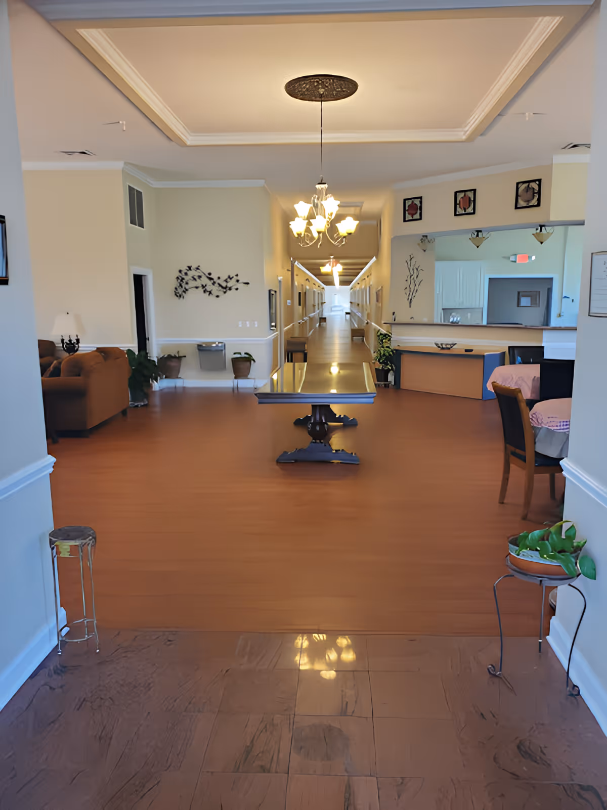 Interior view of a senior care facility lobby with a central table, seating areas, chandeliers, and a long corridor leading to rooms.