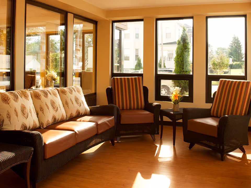 A cozy seating area with a wicker sofa and two wicker armchairs, all with cushions. The sofa has leaf-patterned cushions, and the armchairs have striped cushions. A small table with a flower arrangement sits between the armchairs. Large windows let in natural light and show a view of trees and buildings outside.