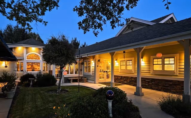 Evening view of a warmly lit single-story building with a covered walkway, surrounded by a well-maintained garden with trees and shrubs.