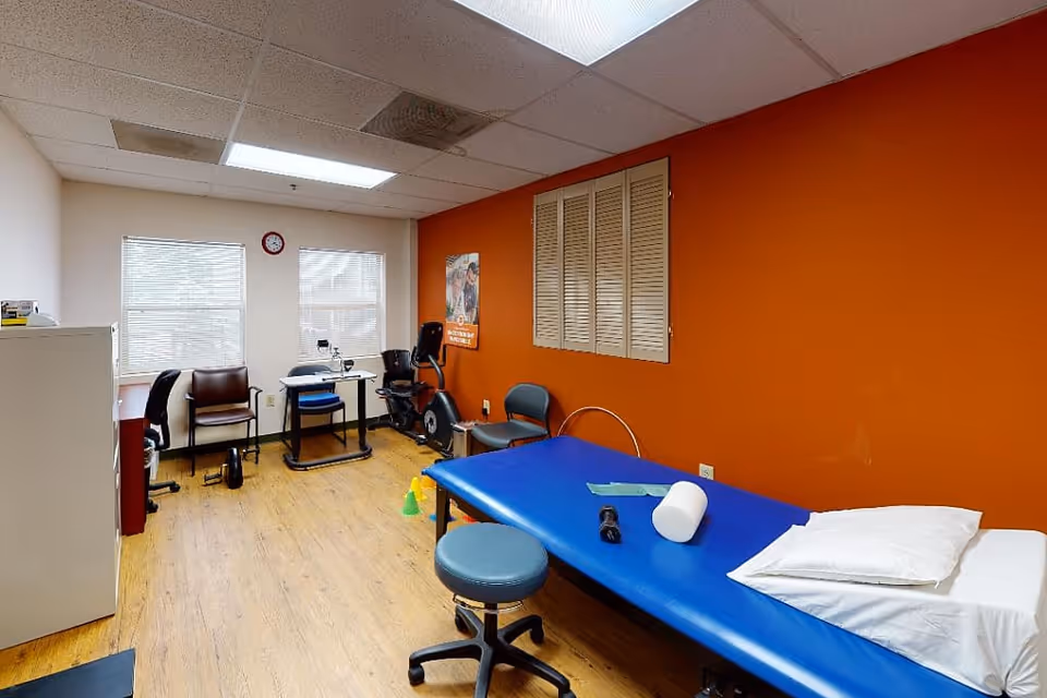 A therapy or rehabilitation room with a blue treatment table covered with a white pillow and a foam roller. There is a rolling stool in front of the table. The room has wooden flooring, two windows with blinds, a red clock on the wall, and an orange accent wall with a poster. There are several chairs, a stationary exercise bike, and some small cones on the floor.