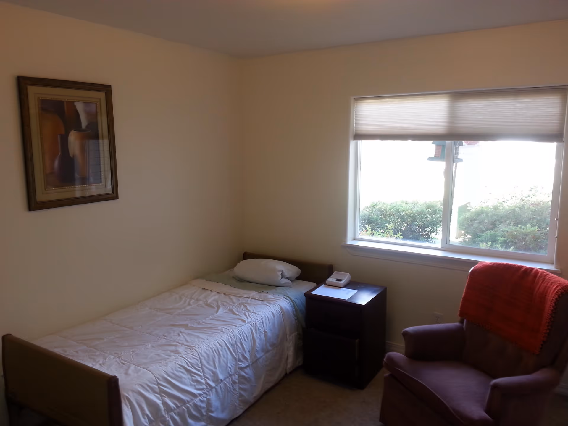 A small bedroom with a single bed covered in a white comforter and a pillow. Next to the bed is a dark wooden nightstand with a white device and a piece of paper on it. There is a window with a partially closed blind letting in natural light, and a purple armchair with a red blanket draped over the back. A framed picture hangs on the beige wall above the bed.