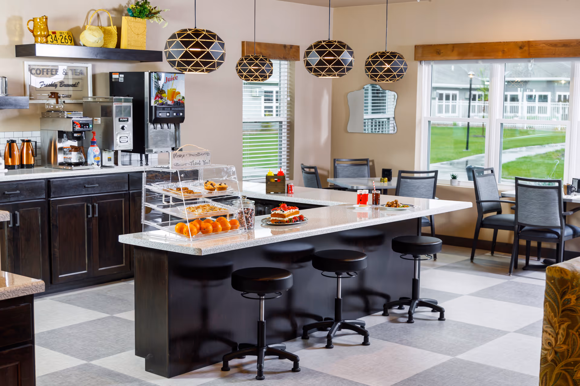Bright communal dining and kitchenette area with a central island and stools, pendant lights, a coffee station, and windows overlooking the grounds.