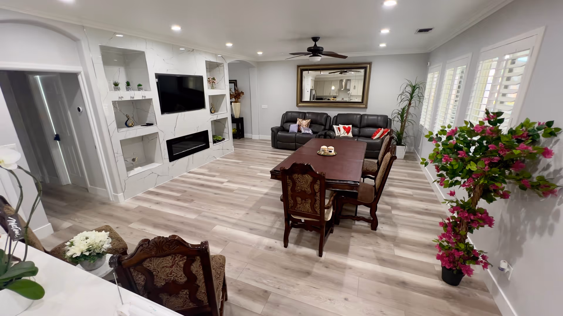 A spacious and modern living room with light wood flooring, a dark wood dining table with six ornate chairs, two black leather sofas with decorative pillows, a large wall mirror, a ceiling fan, and a built-in entertainment center with a flat-screen TV and electric fireplace. There are several plants, including a tall potted plant with pink flowers near the window with white shutters.