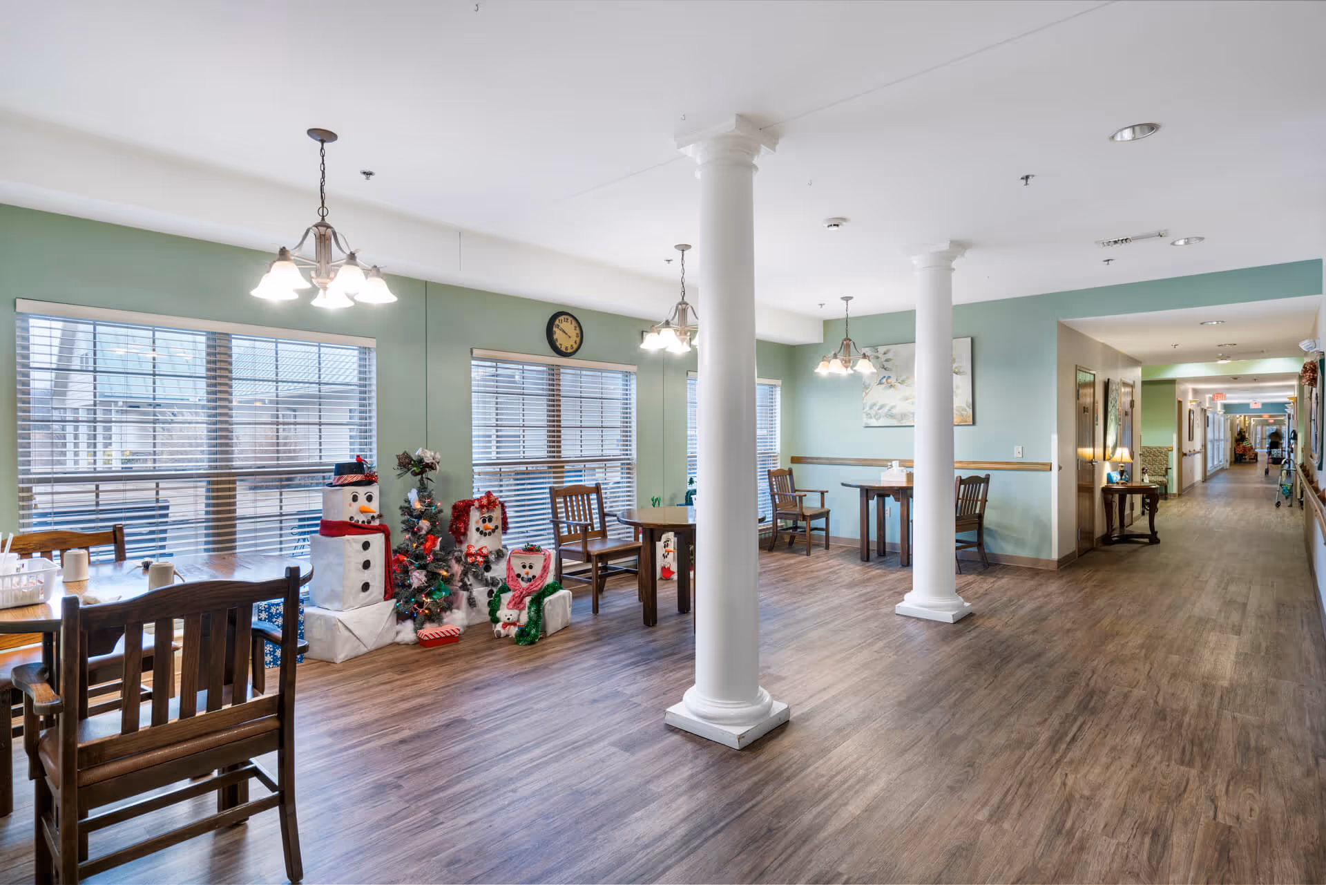 Open communal dining area with wooden tables and chairs, white columns, large windows, and holiday decorations including snowmen and a small tree.