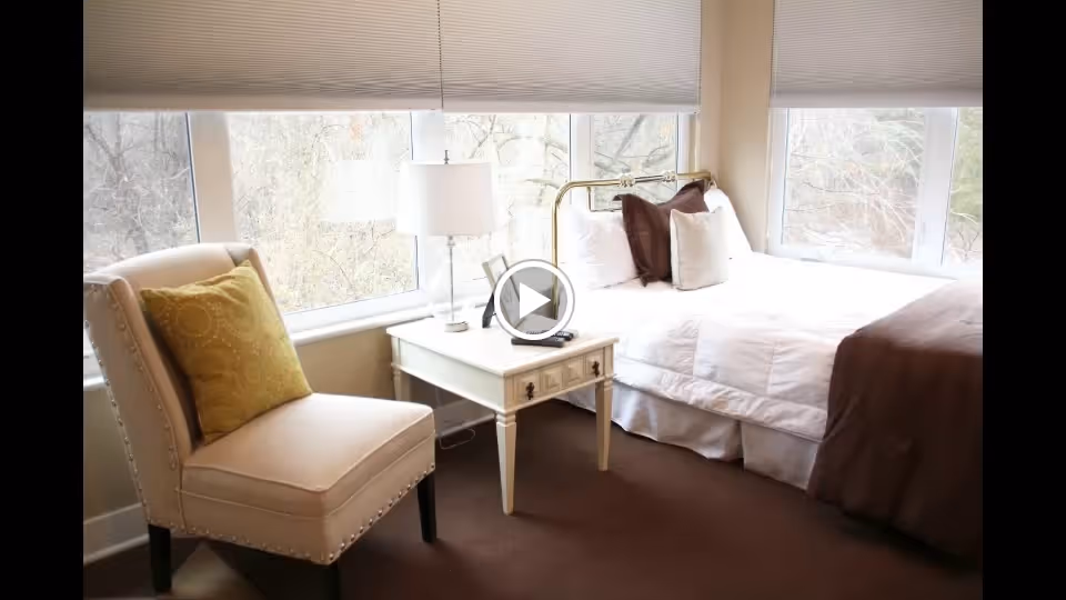 A bright bedroom with large windows showing trees outside. The room features a white bed with brown and white pillows and a brown blanket, a white nightstand with a lamp, phone, and remote control, and a beige upholstered chair with a yellow decorative pillow.