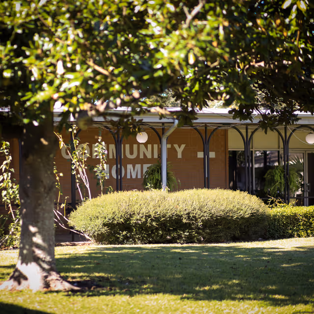 View of the exterior of a building partially obscured by trees and bushes. The building has a sign that reads 'Community Home' and features a covered walkway with hanging plants and round light fixtures.