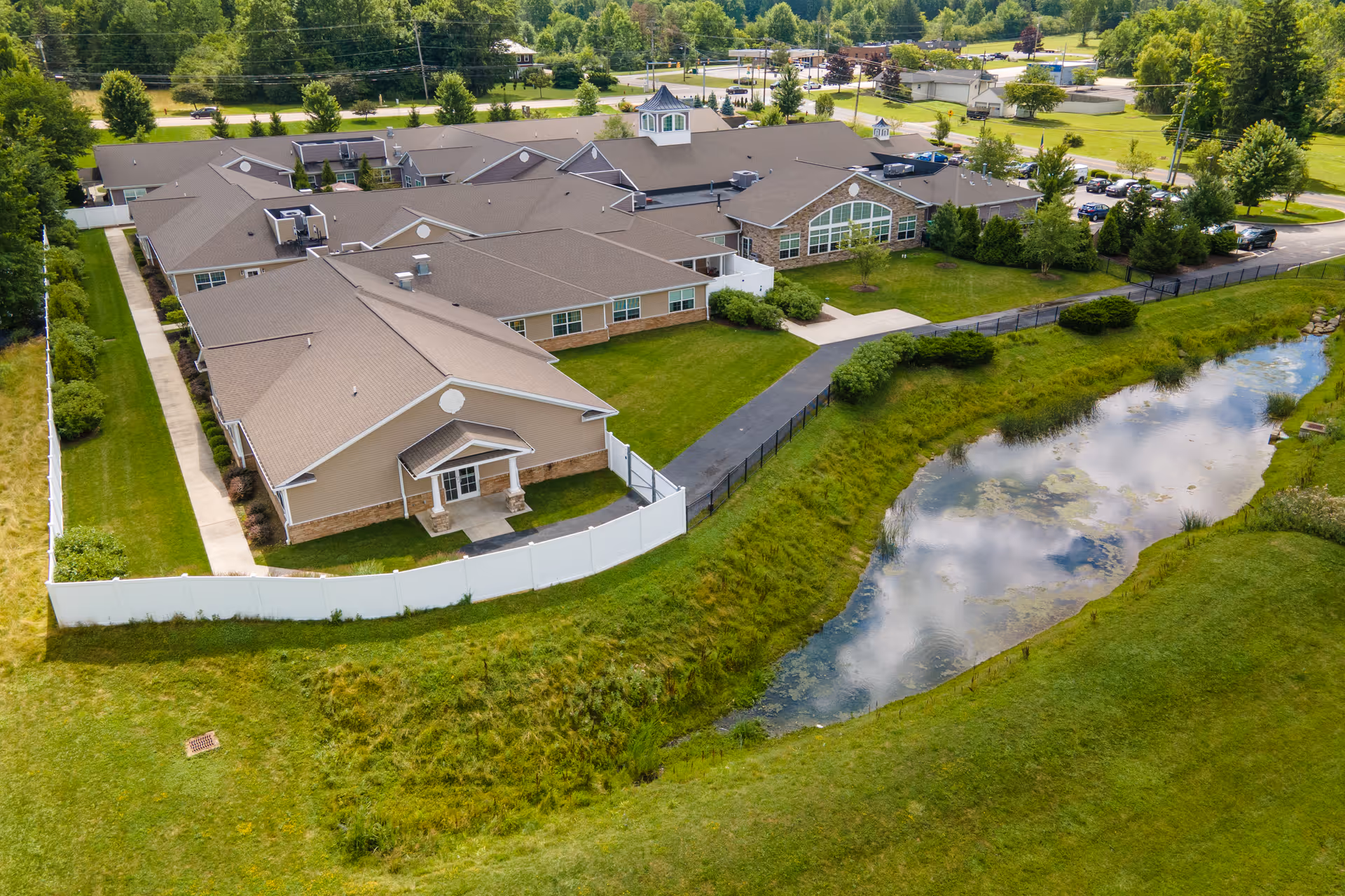 Aerial view of Renaissance of Richfield/Bath senior living facility showing multiple connected buildings with beige siding and brown roofs, surrounded by green lawns, trees, a small pond, and a paved walkway. The facility is enclosed partially by a white fence and has a parking area in the background.