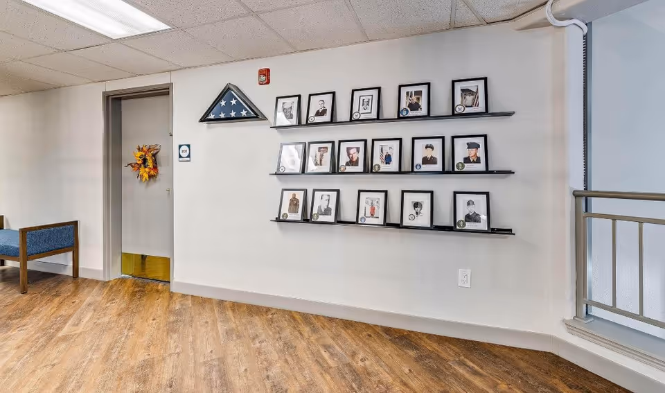 Interior hallway of a senior living facility with a door decorated with a fall-themed wreath, a display of framed photographs and medals on three shelves mounted on the wall, and a folded American flag in a triangular case above the photos. The floor is wood, and there is a railing on the right side.