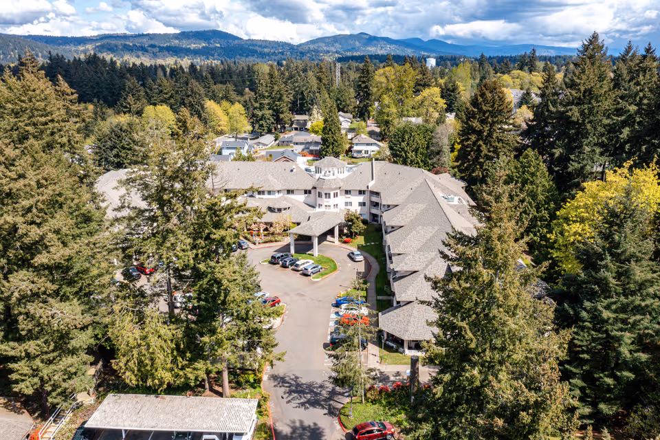 Aerial view of a senior living facility with a covered entrance, parking lot, and surrounding trees and hills.