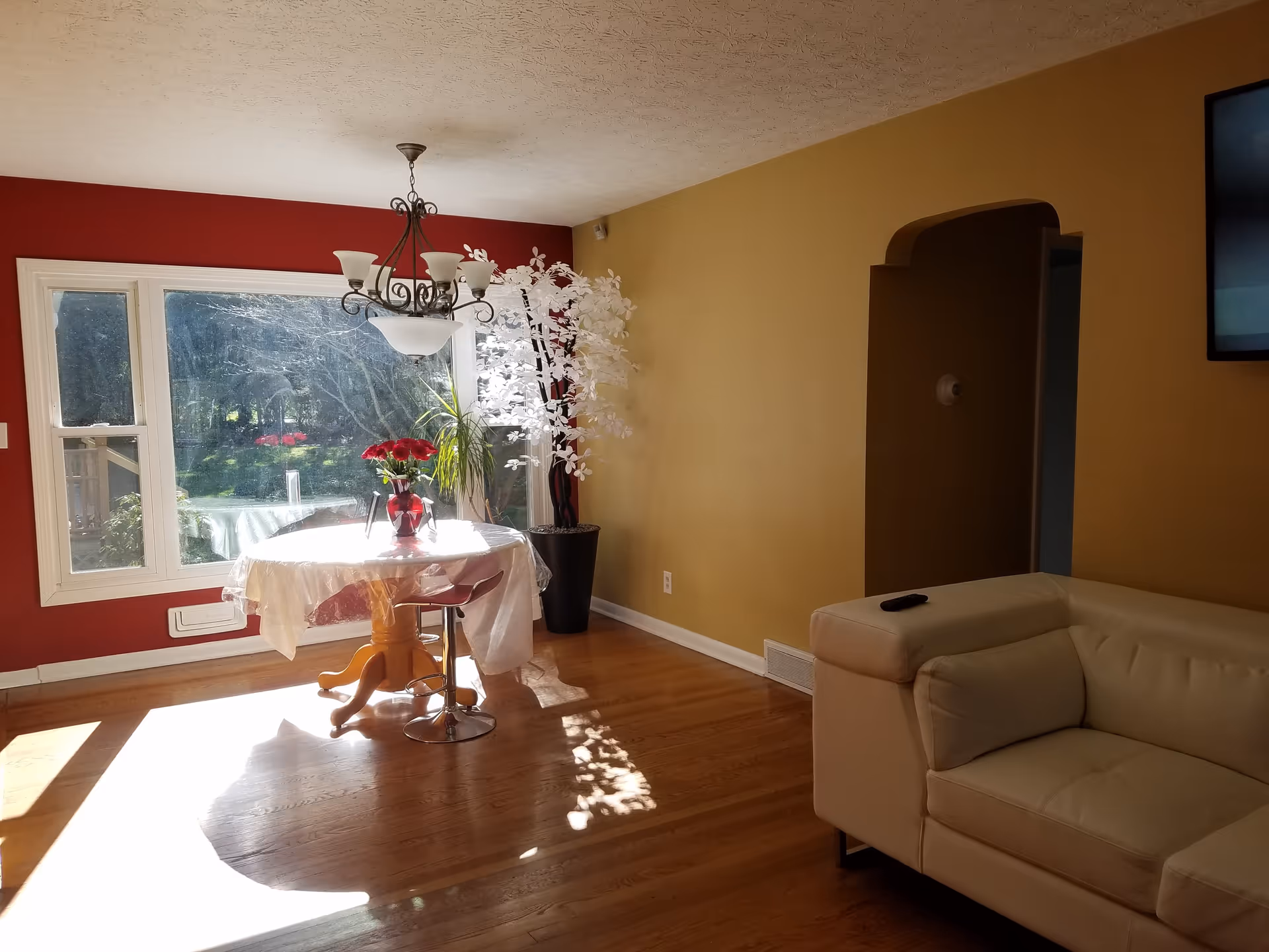 A bright room with wooden flooring featuring a small round dining table covered with a white tablecloth and a vase of red flowers on top. Behind the table is a large window letting in sunlight, with a red accent wall surrounding it. To the right is a beige leather sofa with a remote control on the armrest, a mustard yellow wall with an arched doorway, and a mounted flat-screen TV partially visible.