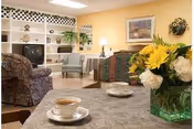 A cozy living room area with comfortable upholstered chairs and sofas arranged around a fireplace and a television. The walls are painted a warm yellow, decorated with framed artwork and a floral wall hanging. A table in the foreground holds a vase with yellow and white flowers and a cup of tea on a saucer.