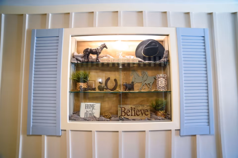 Built-in wall display with blue shutters containing decorative horse figurines, a cowboy hat, potted plants, and signs reading 'Believe' and 'Home'.