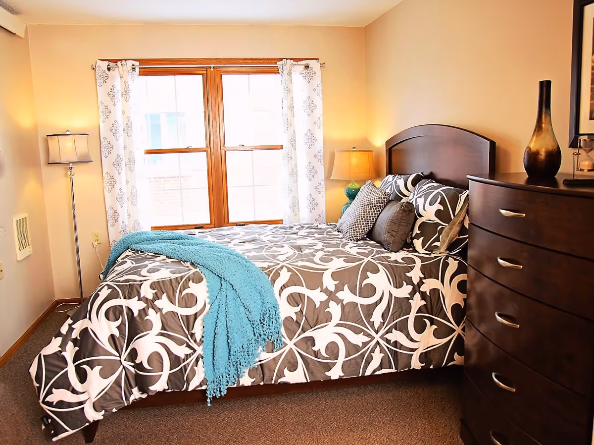 A cozy bedroom with a bed covered in a gray and white patterned comforter, accented with multiple pillows and a turquoise throw blanket. The room features a wooden headboard, a matching dark wooden dresser with a decorative vase on top, two lamps providing warm lighting, and a window with white patterned curtains letting in natural light.