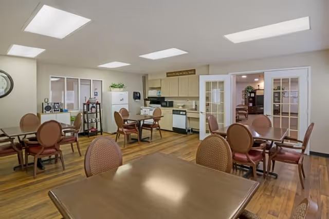 A well-lit dining area with multiple wooden tables and cushioned chairs arranged neatly. The room features a kitchen area with cabinets, a refrigerator, microwave, and dishwasher. There are glass double doors leading to another room, and the floor is wooden. The ceiling has several rectangular recessed lights.