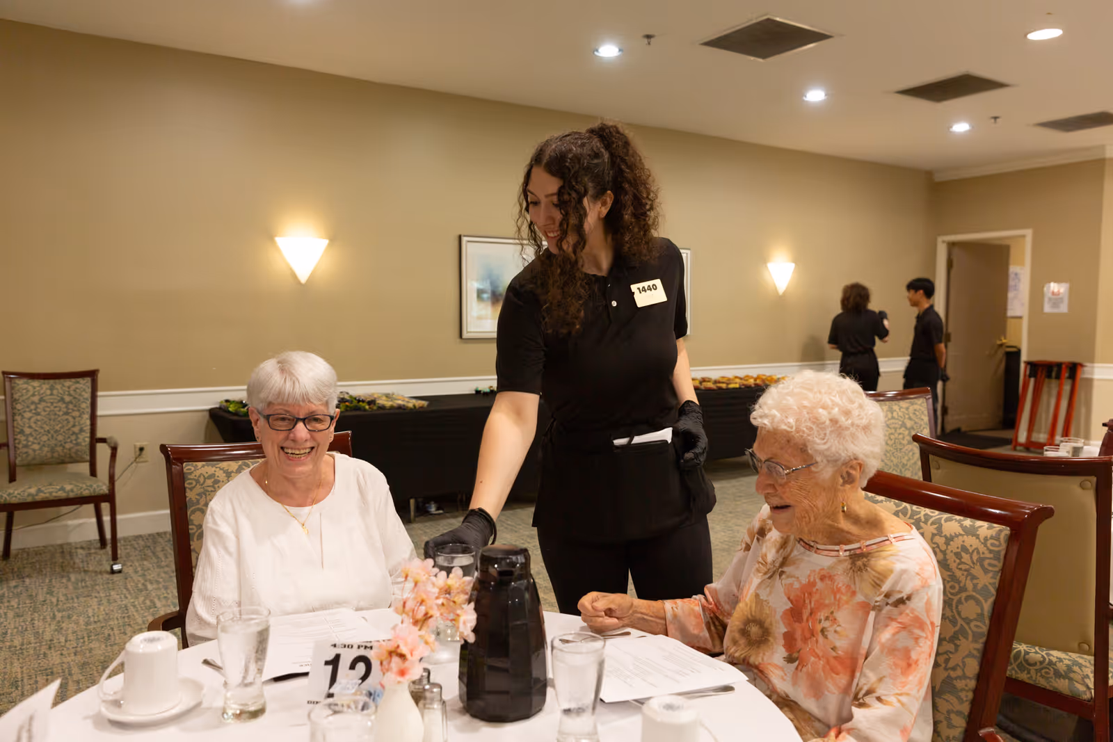Two elderly women sitting at a round dining table in a well-lit room, smiling and interacting with a young female server who is pouring a drink. The table has menus, glasses, a coffee pot, and a small flower arrangement. In the background, two other staff members are seen near a doorway.