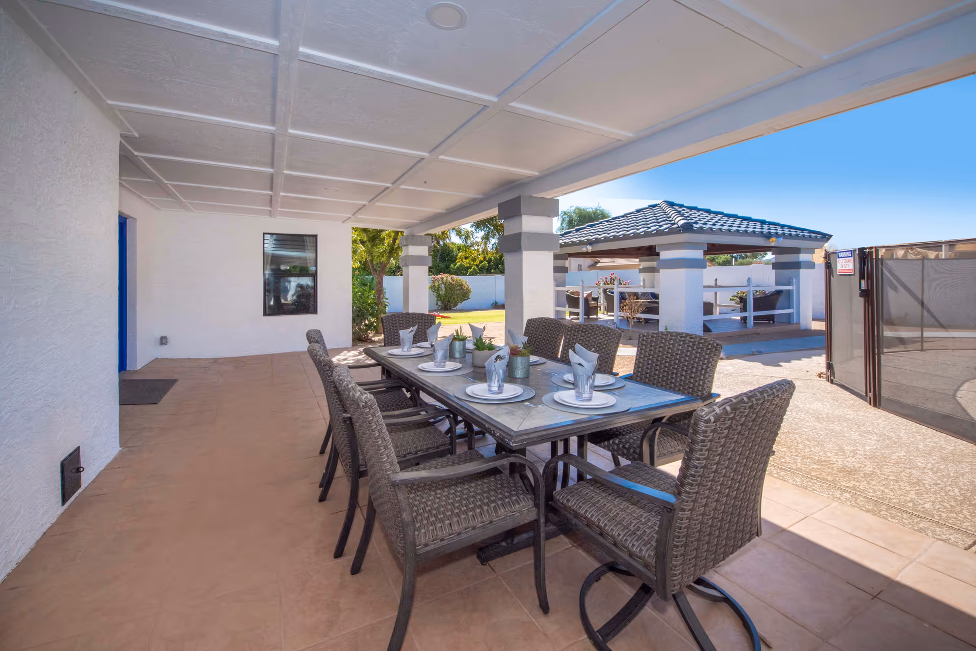 Outdoor covered patio area with a dining table set for six people with plates, napkins, and cups. The patio has a tiled floor and white ceiling with beams. In the background, there is a gazebo with seating and a white fence enclosing the area under a clear blue sky.