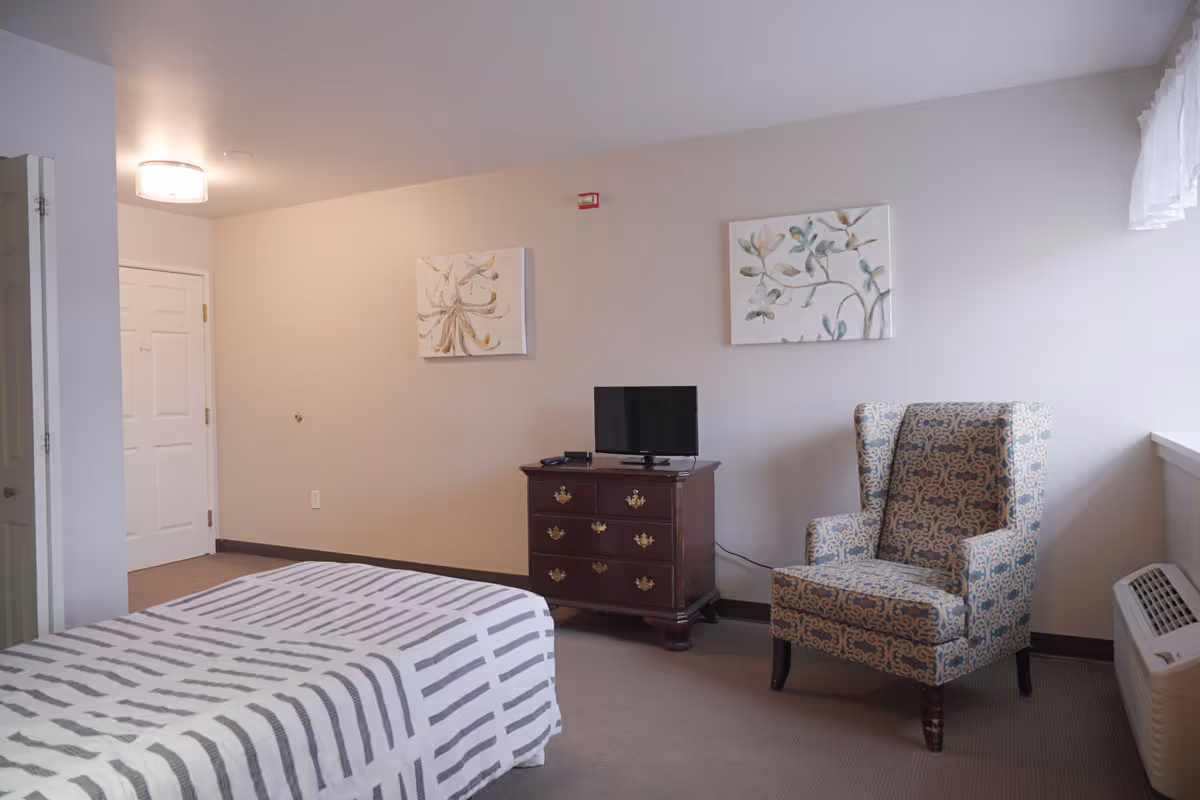 A senior living facility bedroom with a bed covered in a striped blanket, a wooden dresser with a small flat-screen TV on top, a patterned armchair, two floral paintings on the wall, a window with sheer curtains, and a white door in the background.